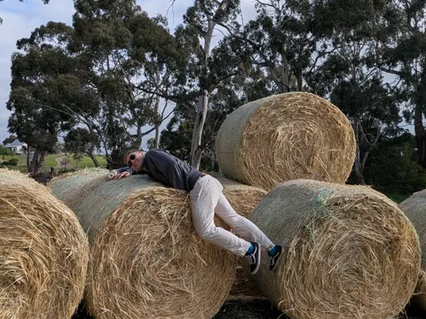 A man lying draped across a hay bale, sunglasses on, arm hanging down.